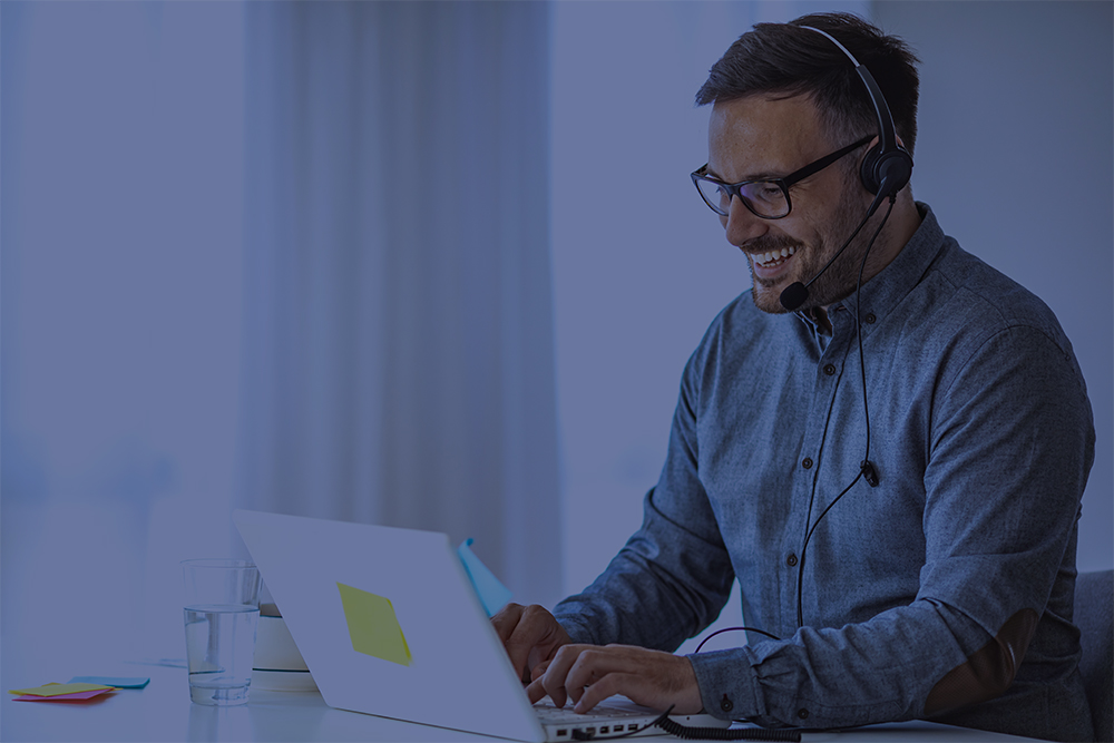 Homem sorridente usando óculos e fones de ouvido com microfone, sentado em uma mesa branca enquanto digita em um notebook com um post-it amarelo colado. Ao lado, há um copo de água e alguns post-its coloridos. A cena sugere uma videochamada ou atendimento remoto em ambiente de trabalho.
